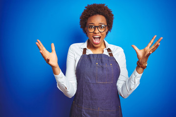 Sticker - Young african american woman shop owner wearing business apron over blue background celebrating crazy and amazed for success with arms raised and open eyes screaming excited. Winner concept