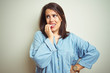 © Krakenimages.com - Young beautiful brunette woman wearing casual blue denim shirt over isolated background looking stressed and nervous with hands on mouth biting nails. Anxiety problem.