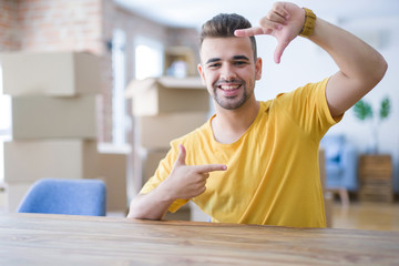 Sticker - Young man sitting on the table with cardboard boxes behind him moving to new home smiling making frame with hands and fingers with happy face. Creativity and photography concept.