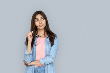 Curious young adult indian woman standing isolated on grey background with copy space. Dreaming girl in denim wear looking away holding glasses in hand uncertain with question interested or doubting