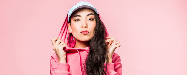 panoramic shot of attractive asian woman in pink pullover and cap looking at camera isolated on pink