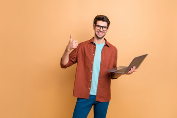 Portrait of his he nice attractive cheerful cheery successful content brunet guy holding in hands laptop showing thumbup isolated over beige color pastel background