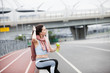 Athletic woman in sports outfit after jogging, drinks water from bright green bottle, sitting on the parapet of bridge.