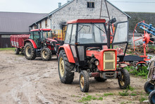 Red Tractors And Agricultural Equipment In The Yard Of A Dairy Farm. Cowshed In The Background. Podlasie, Poland.