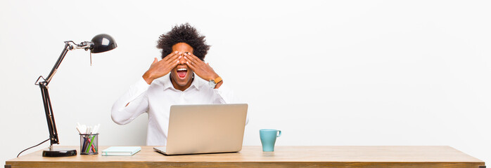 Wall Mural - young black businessman smiling and feeling happy, covering eyes with both hands and waiting for unbelievable surprise on a desk