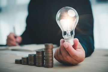 businessman hand holding lightbulb with coins stacking on desk.saving energy and money concept.