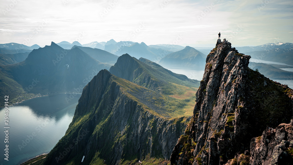 Adventurous man is standing on top of the mountain and enjoying the beautiful view during a vibrant sunset. Taken on top Senja, Norway  - obrazy, fototapety, plakaty 