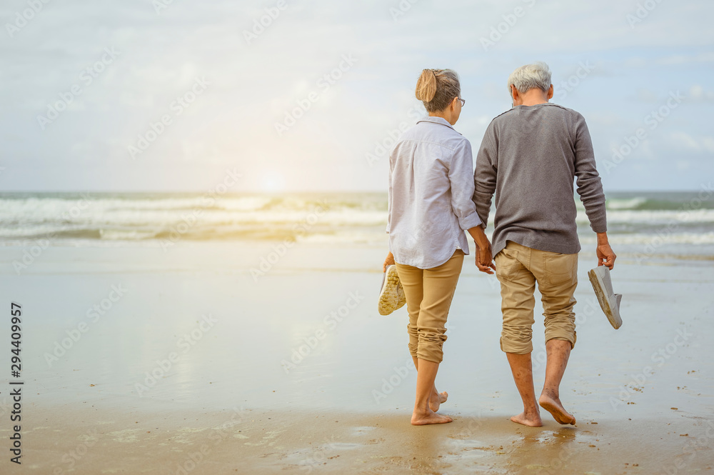 Senior couple walking on the beach holding hands at sunrise, plan life insurance at retirement concept. - obrazy, fototapety, plakaty 