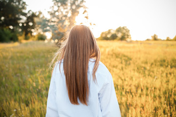 Beautiful young woman in white shirt walking outdoors at sunset. Back