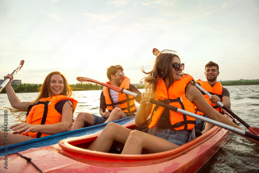 Happy young caucasian group of friends kayaking on river with sunset in the backgrounds. Having fun in leisure activity. Happy male and female model laughting on the kayak. Sport, relations concept. - obrazy, fototapety, plakaty 