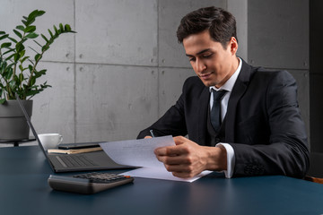 Friendly young businessman in dark suit working with documents at loft office table with laptop and calculator. Concept of legal work and startup