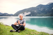 A Senior Man Pensioner Sitting By Lake In Nature, Doing Yoga Exercise.