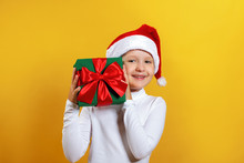 Happy Little Girl In Santa Hat Holds Christmas Gift. A Child In A White Turtleneck On A Yellow Background