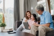 © tirachard - Asian grandparents and granddaughter using tablet at home. Senior Chinese, grandpa and grandma happy spend family time relax with young girl checking social media, lying on sofa in living room concept