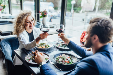 Romantic pretty young couple at restaurant raising a toast. Beautiful couple with glasses of red wine in restaurant. Couple toasting wine glasses during a romantic dinner in a gourmet restaurant.