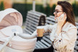 Smiling mother with a newborn baby in a stroller drinks tea or coffee in a street cafe and talking by her phone. Fashionable modern mom with a baby.