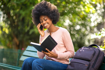 Wall Mural - happy smiling black woman sitting outside with book bag and talking with mobile phone