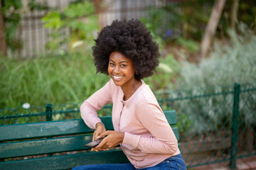 Wall Mural - young african american woman sitting on park bench with phone in hand