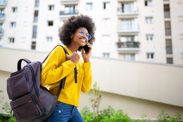 Poster - Side of smiling african american woman with glasses and bag walking and talking with cellphone in city