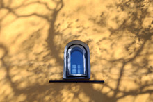 Single Arched Window On A Yellow Wall With Shadows From Tree Branches. Blue Sky Reflected In The Window Pane. In Tallinn Estonia.