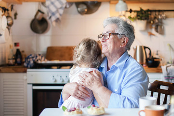Happy senior woman is hugging child in cozy home kitchen. Grandmother and cute little girl are smiling. Kid is enjoying kindness, warm hands, care, support. Family is cooking together.