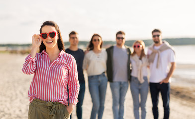 friendship, valentine's day and people concept - happy woman in heart-shaped sunglasses with group of friends on beach in summer