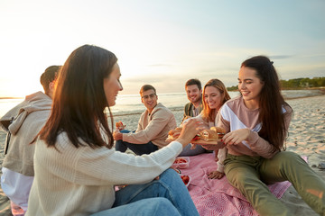 Poster - friendship, leisure and fast food concept - group of happy friends eating sandwiches or burgers at picnic on beach in summer