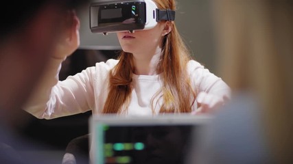 Camera tilting up from laptop with code to young woman exploring virtual reality near developers during VR app test in office