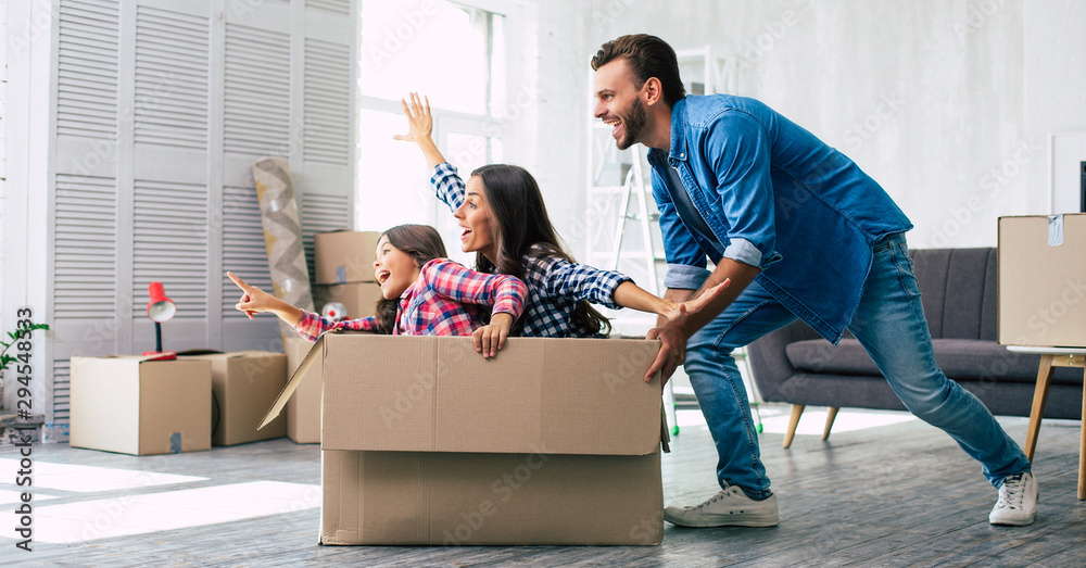 Little girl is riding in a cardboard box and pointing straight ahead while her mother imitates a plane and laughs, and father is pushing the box, enjoying their time together in their new house. - obrazy, fototapety, plakaty 