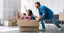 Little Girl Is Riding In A Cardboard Box And Pointing Straight Ahead While Her Mother Imitates A Plane And Laughs, And Father Is Pushing The Box, Enjoying Their Time Together In Their New House.
