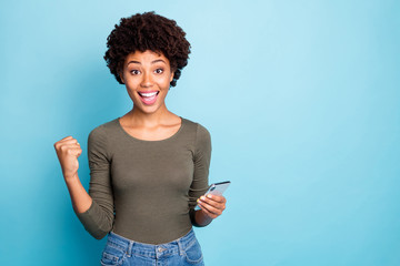 Yes more followers. Portrait of cheerful curly hair girl hold use smartphone get notification celebrate lottery victory raise fist scream wear green pullover isolated blue color background