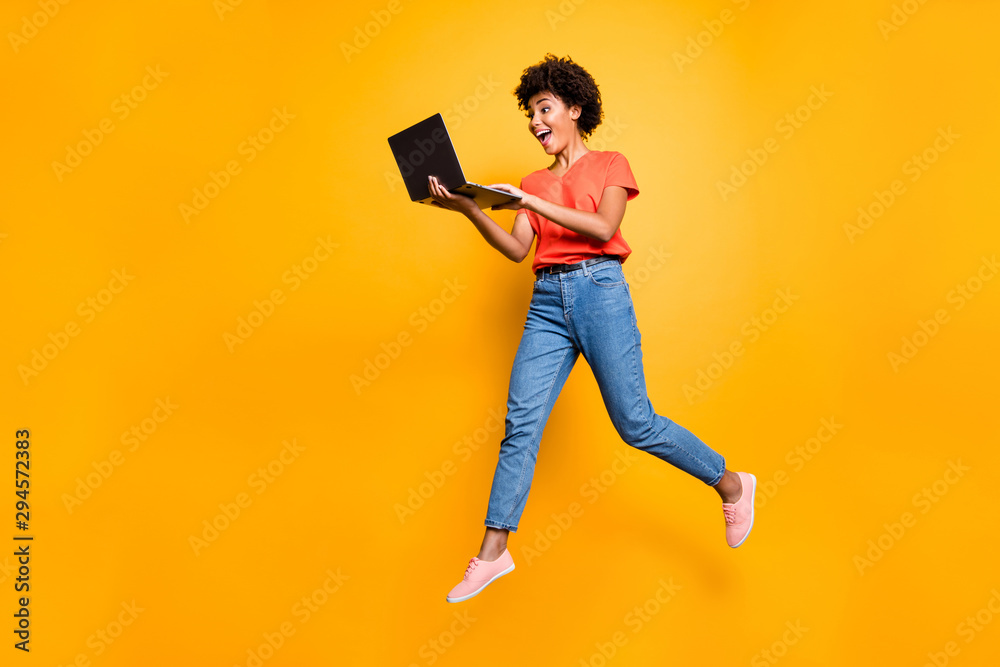 Full length photo of excited funny funky curly hair girl use her laptop find news information about sales jump wear denim jeans stylish trendy sneakers t-shirt isolated over yellow color background - obrazy, fototapety, plakaty 