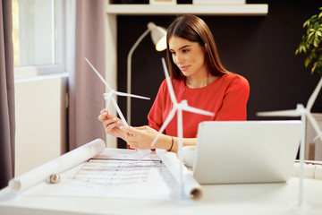 Dedicated hardworking pretty businesswoman dressed in formal wear sitting in office and holding windmill model. On table are blueprints and laptop. Sustainable development concept.