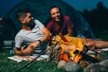 Wall Mural - close up of campers barbecue sausages. happy young couple on night camping by the camping fire