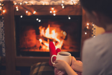 Close up cup of hot tea in woman's hands sitting near fireplace with festive Christmas lights in cozy room.