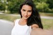 © iuricazac - Close-up portrait of a beautiful young brunette smiling woman taking self portrait on nature background in the park. Pretty female making selfie on her device outdoors. People, lifesetyle and travel