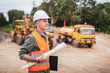 © Pituk - Caucasian handsome engineer is stand and smile holding a laptop with roll of paper on road construction vehicle background.
