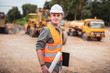 © Pituk - Caucasian handsome engineer is stand and smile holding a laptop with roll of paper on road construction vehicle background.