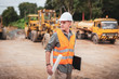 © Pituk - Caucasian handsome engineer is holding a laptop with roll of paper on road construction vehicle background. Engineer work concept