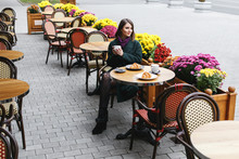 Beautiful Young Girl Wearing Green Coat Sitting At A Table In Cozy Street Outdoor Cafe And Drinking Coffee With A Croissant. Restaurant Terrace Is Decorated With Chrysanthemum Flowers Bushes In Autumn