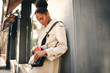 © Anton - Side view of beautiful African American girl in stylish trench coat taking phone out of bag outdoor