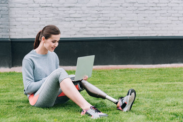 smiling disabled sportswoman using laptop while sitting on grass