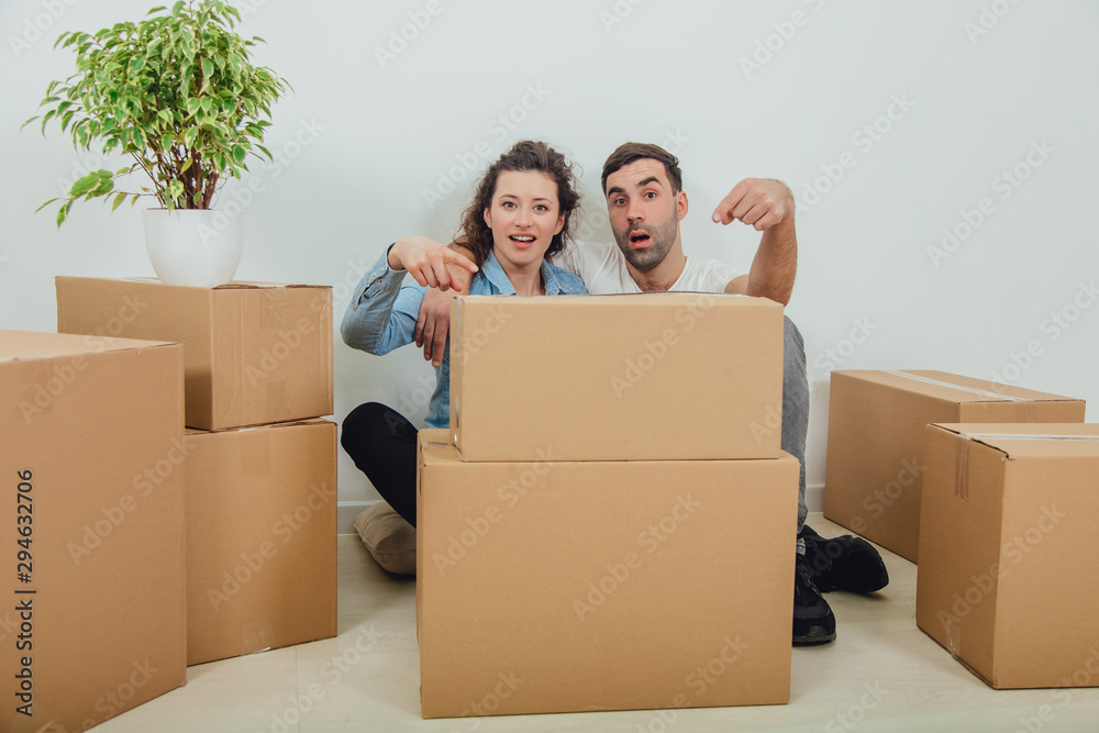Funny wife and husband playing around, sitting behind the big carton boxes, pointing fingers at them, looking foolishly. - obrazy, fototapety, plakaty 