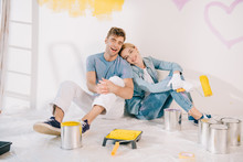 Happy Young Woman Holding Yellow Paint Roller While Sitting On Floor Near Boyfriend