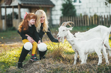 Blonde Woman With Her Red-heired Little Daughter Play With White Goat On Nature. Love And Care. Village Animals.