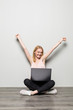 © dianagrytsku - Attractive woman holding laptop computer while sitting on the floor with legs crossed and looking away at copy space isolated over gray background