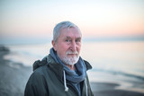 Eldery man with gray hair and beard with blue eyes in green coat on in the beach at sunrise