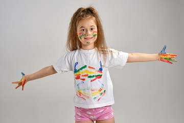 Wall Mural - Little girl in white painted t-shirt is posing standing isolated on white and gesticulating with her colored in different paints palms and face. Art studio. Close-up.