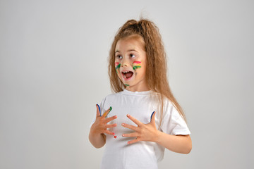 Wall Mural - Little girl in white painted t-shirt is posing standing isolated on white and gesticulating with her colored in different paints palms and face. Art studio. Close-up.