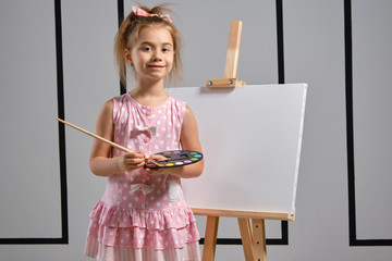 Wall Mural - Little girl artist in a pink dress is standing behind easel and painting with brush on canvas at art studio with white walls. Medium close-up shot.